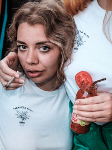 A woman holding a glass of tomato sauce with a straw in it is offering a cigarette to a woman sitting next to her. Both a wearing white "Paradeis Kush" shirts.