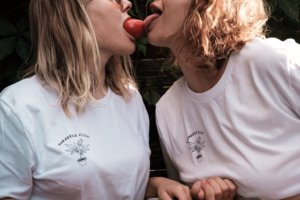 A woman wearing a white "Paradeis Kush" shirt is holding a tomato in her mouth. Another woman, wearing the same shirt, is licking the tomato.