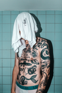 A heavily tattooed man is standing in front of a turquoise tiled wall. He is wearing a white "Onlyfans" shirt over his face.