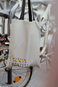 A beige "Prada-Prater" bag is hanging on the handle of an old bicycle. "Prada" is crossed out with a yellow stroke. The bike has the same beige and yellow colors on its' frame.