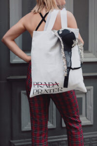 A woman is standing in front of a grey door. She is wearing red checkered pants and is holding a white "Prada-Prater" bag. "Prada" is crossed out with a red stroke. A black bra is peeking out of the bag.