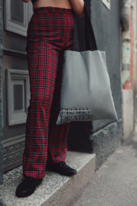 A woman is standing in front of a grey door. She is wearing red checkered pants and is holding a grey "Prada-Prater" bag. "Prada" is crossed out with a white stroke.