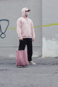 A man with a pastel pink hoodie, mask, cap and shoes is holding a dark pink "Prada-Prater" bag. "Prada" is crossed out with a pastel pink stroke. He is standing in the street with a grey wakl in the back.