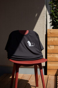 The black shirt for "Container 25" stretched over a red chair. It shows the "Container 25" logo, which is two abstract houses, leaning away from each other. It is shown from the front.