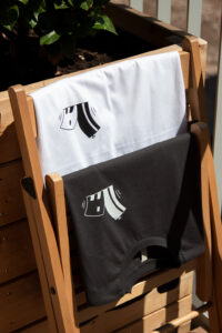 A black and a white "Container 25" shirt, both with the logo, which shows two abstract houses, leaning away from each other, in the opposite color respectively. They are hanging over a closed folding chair.