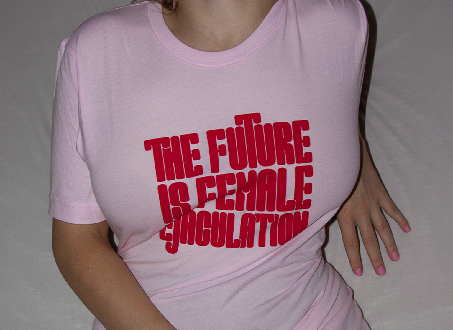 A Woman, sitting on a bed, wearing the shirt in pink with red text. Her face is cropped.