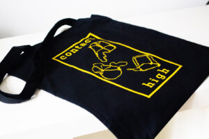 A black bag with the bright yellow "Contact High" logo, showing their logotype, abstract champagne muselets and a bold border around it all. It is shown from a medium angle.