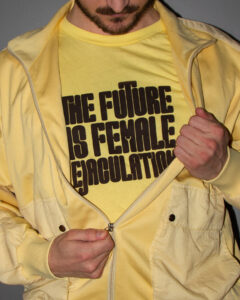 A man, zipping down his jacket, wearing the yellow shirt with the brown text underneath. His face is cropped